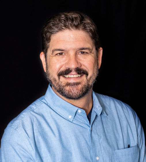 Brandon Bernard headshot- smiling white man with brown hair and beard, wearing a blue oxford shirt